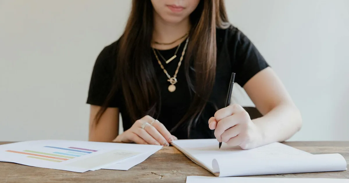 A woman writing at a wooden table, surrounded by papers in a bright indoor setting, ideal for productivity and focus.