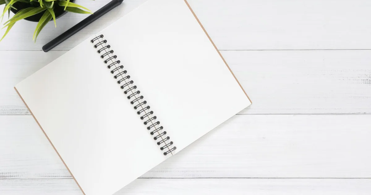Open spiral notebook on a white desk with a black pen and a green plant, ideal for planning and organization.